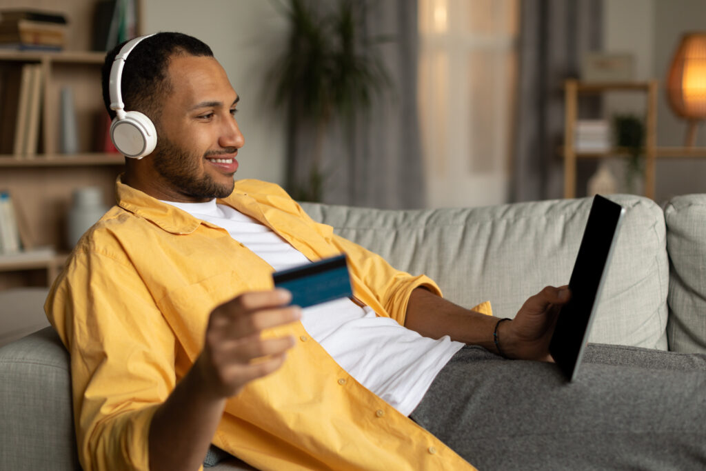 Hombre alegre con portátil y tarjeta de crédito tumbado en el sofá, con auriculares, comprando por Internet en casa.