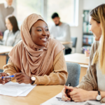 Estudiante musulmana feliz hablando con su compañera de clase, ambas sentadas en un pupitre.